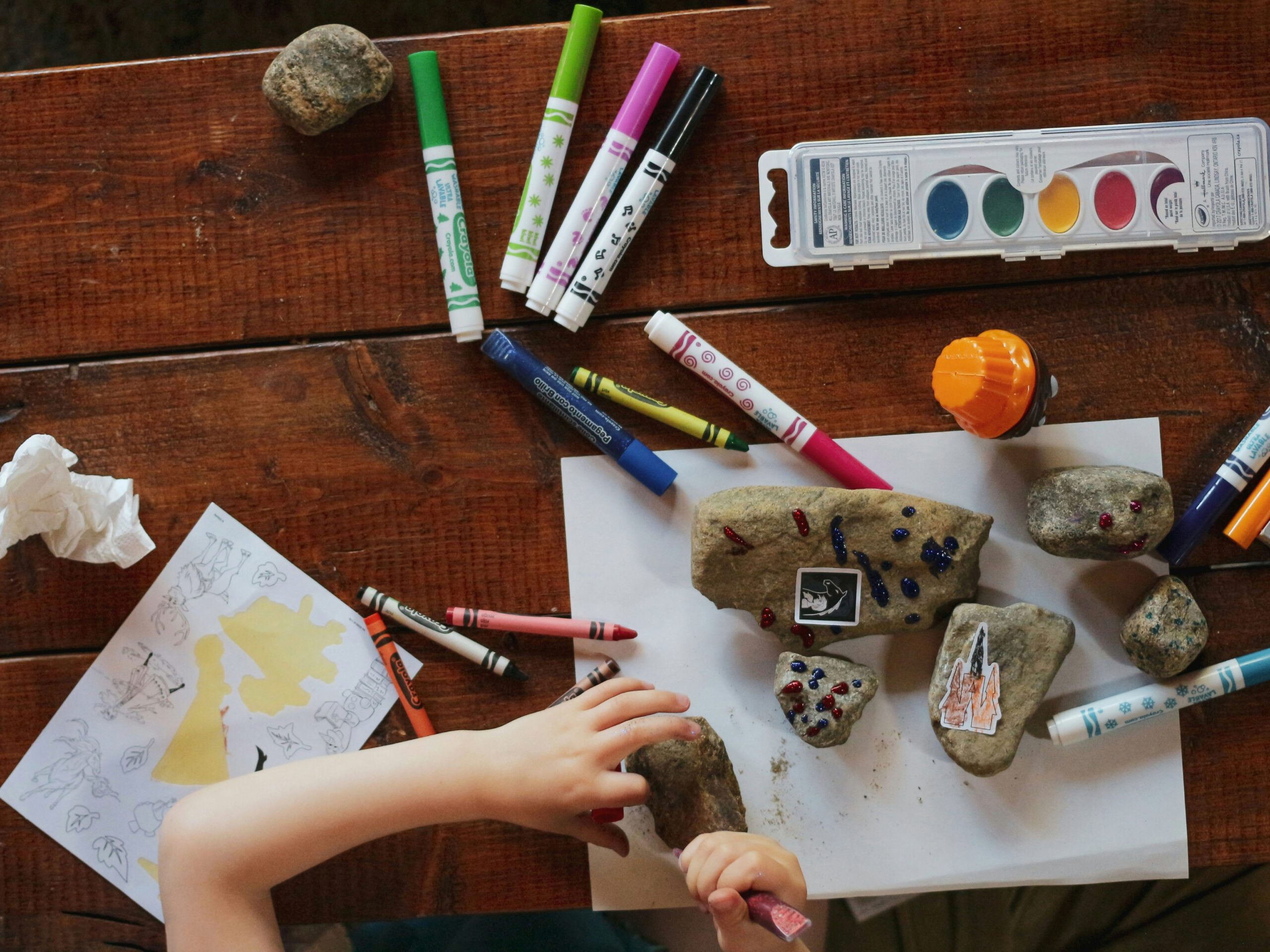 Markers, paint on table with a child's hands coloring rocks